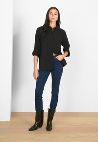 Chemise noire à boutons avec des manches roulées, associée à un jean bleu ajusté et des bottes de cowboy noires, sur fond blanc uni.