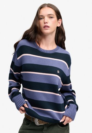 Jonge vrouw die een blauw, zwart en lichtroze gestreepte trui met lange mouwen draagt, en de zoom van de trui met beide handen vasthoudt.