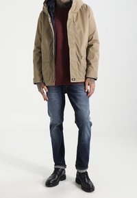 Beżowa kurtka z kapturem z ciemną podszewką, pod spodem brązowy sweter, niebieskie dżinsy i czarne buty. Posiada kieszenie i swobodny krój.