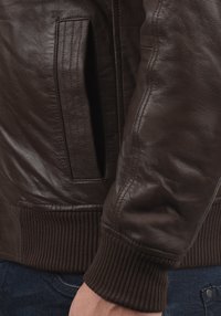 Dettaglio delle maniche e della tasca di una giacca di pelle marrone con polsini e vita a coste, indossata sopra jeans di denim blu.