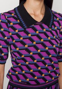 Dzianinowa bluzka z wyrazistym geometrycznym wzorem w kolorach fioletowym, czarnym, pomarańczowym i niebieskim. Czarny kołnierz i krótkie rękawy. Miękki, teksturowany materiał.