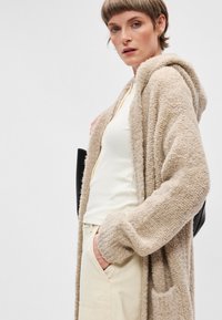 Cardigan beige fluffy avec capuche, épaules tombantes et grandes poches, porté sur un haut ajusté jaune pâle et un pantalon crème.