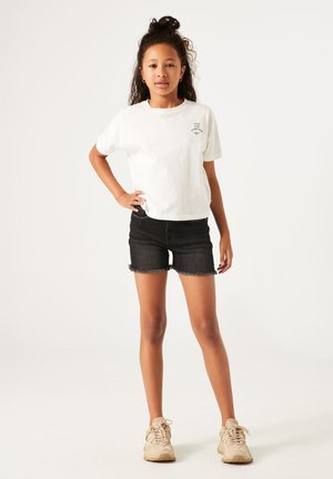 T-shirt en coton blanc avec un design imprimé sur le devant, associé à un short en denim noir avec des bords effilochés. Des baskets beiges complètent la tenue.