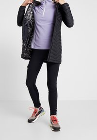 Persoon draagt een zwarte doorgestikte jas met glanzende voering, een lavendelkleurig vest met rits, zwarte legging en grijze sneakers met oranje veters.