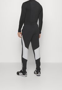 Schwarzes Sportoutfit mit langen Ärmeln und eng zulaufenden Hosen mit grauen Akzenten. Der Stoff wirkt glatt, dehnbar und eng anliegend. Schwarze Schuhe mit weißen Streifen.