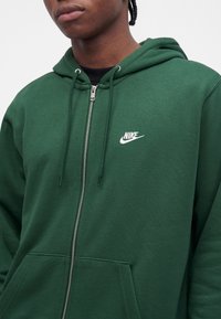 Άνδρας που φοράει σκούρο πράσινο φούτερ Nike με φερμουάρ και λευκό λογότυπο στο στήθος, μαύρο πουκάμισο από κάτω, σε απλό ανοιχτό φόντο.