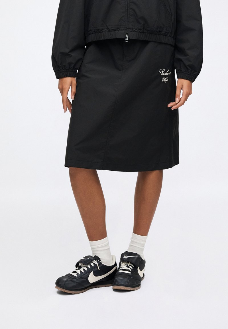 Black, knee-length skirt made of lightweight material, featuring an elastic waistband and embroidered detail. Paired with black Nike sneakers and white socks.