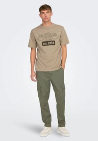 Camiseta de algodón beige con el texto gráfico marrón "Central Perk EST. 1994." Combinada con pantalones cargo verde oliva que cuentan con múltiples bolsillos. Zapatillas blancas.