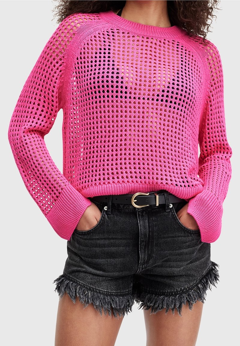 Różowy sweter z dzianiny o luźnym, otwartym splocie oraz ściągaczami na mankietach, w zestawieniu z czarnymi szortami dżinsowymi z postrzępionymi krawędziami i czarnym paskiem.