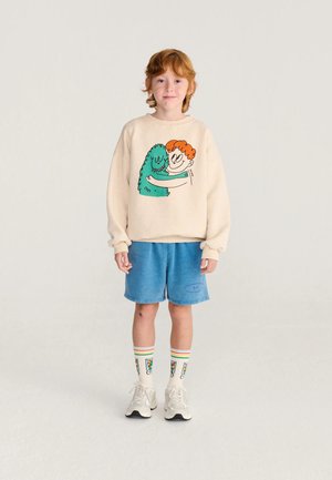 Niño pequeño pelirrojo que lleva una sudadera beige con un diseño de abrazo de dibujos animados, pantalones cortos azules, calcetines coloridos y zapatillas blancas, de pie sobre un suelo blanco.