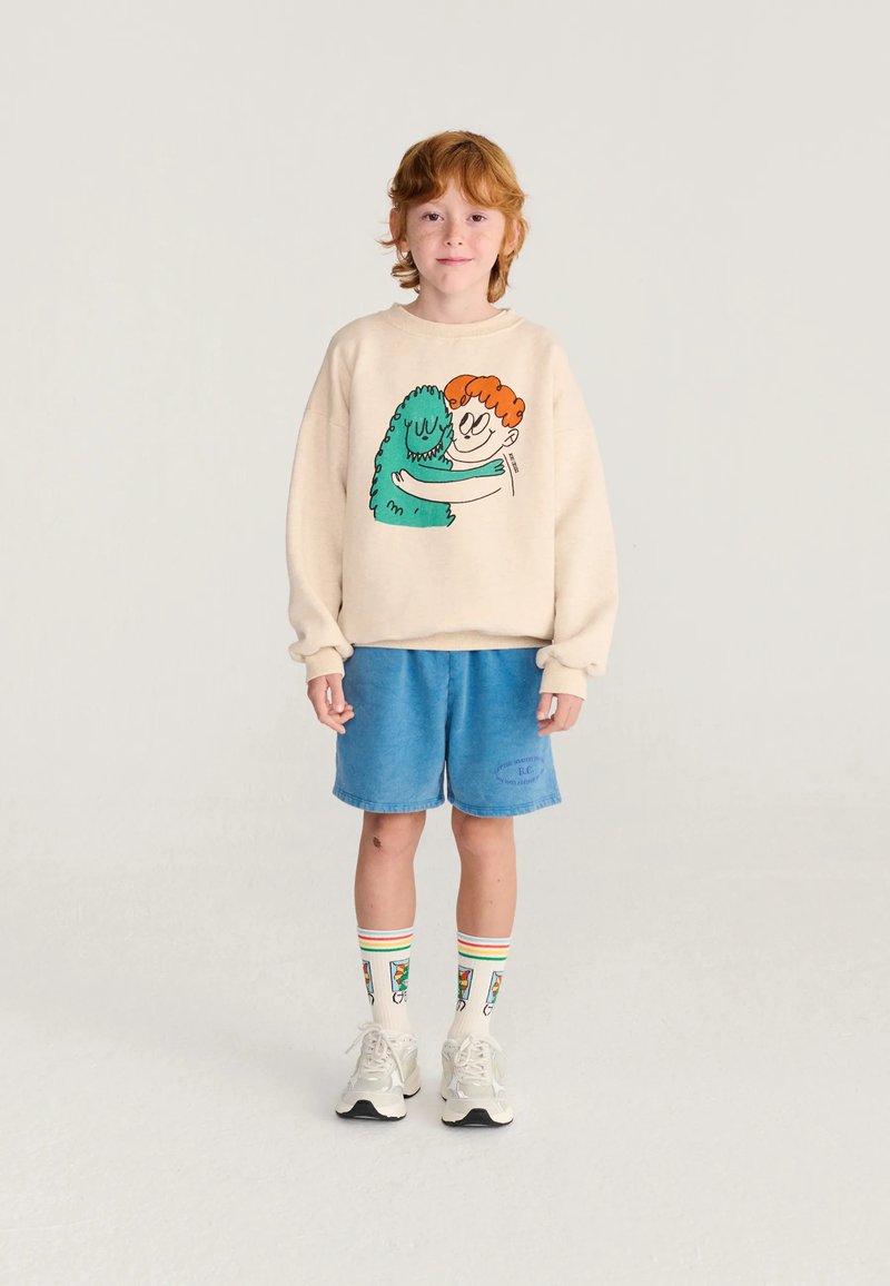 Jeune enfant roux portant un sweat-shirt beige avec un motif de câlin de dessin animé, un short bleu, des chaussettes colorées et des baskets blanches, debout sur un sol blanc.