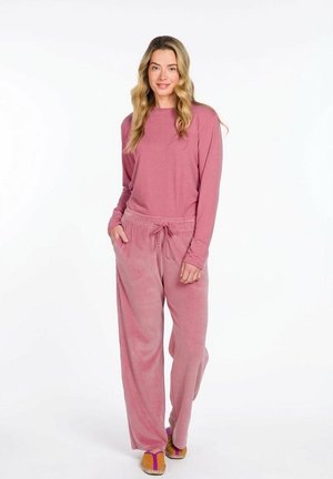 Woman wearing a long-sleeve pink top and loose pink velour pants, standing with hands in pockets on a white background.