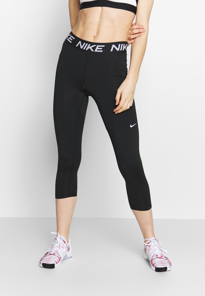 Personne en forme portant des leggings courts Nike noirs et des baskets blanches avec des touches rouges, debout devant un fond clair uni.