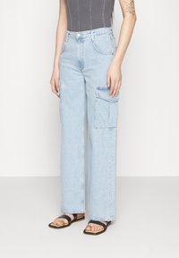 Světle prát široké denní cargo kalhoty s velkými bočními kapsami, nošeny s černými plochými sandály a bezrukávovým šedým žebrovaným topem.