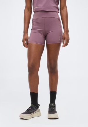 Personne portant un short de sport taille haute mauve, des chaussettes noires mi-mollet et des baskets Adidas grises, debout sur un fond clair uni.