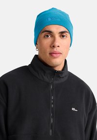 Man in een felblauwe Jack Wolfskin-beanie en een zwarte fleecejack met ritssluiting en logo op de borst, met oorbellen zichtbaar op beide oren.