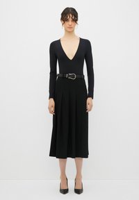 Bodysuit negro de manga larga con escote en V profundo, combinado con una falda midi negra con pliegues y un cinturón negro con hebilla plateada. Tacones negros.