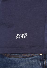 Navy blue t-shirt made of soft fabric, featuring a white logo "BLND" with "SINCE 1993" at the bottom hem, above denim jeans.