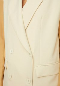 Beige Doppelreiher-Blazer mit Reverskragen, ausgestattet mit cremefarbenen Knöpfen, einer aufgesetzten Tasche und einer glatten, gewebten Textur.
