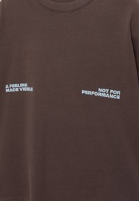 Sötétbarna póló világoskék felirattal: bal oldalon "EGY ÉRZÉS LÁTHATÓVÁ TÉVE", jobb oldalon "NEM TELJESÍTMÉNYRE".