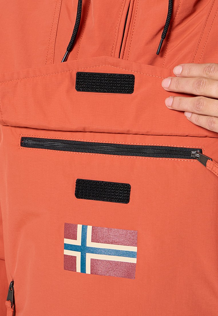 Giacca arancione con una tasca frontale con zip e dettagli in Velcro. Presenta una toppa con la bandiera della Norvegia e lacci al colletto. Materiale liscio e leggero.