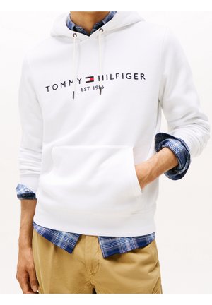 Mężczyzna w białej bluzie z kapturem Tommy Hilfiger na niebieskiej koszuli w kratę i spodniach khaki, z jedną ręką w kieszeni bluzy.