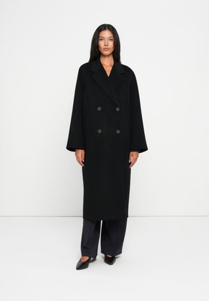 Manteau long noir croisé avec de larges revers et de gros boutons, en tissu texturé, associé à un pantalon sombre et des chaussures noires.