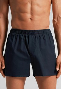 Námořnické modré plavky s elastickým pasem, hladkou texturou a mírně volným střihem. Žádné viditelné vzory ani detaily hardware.