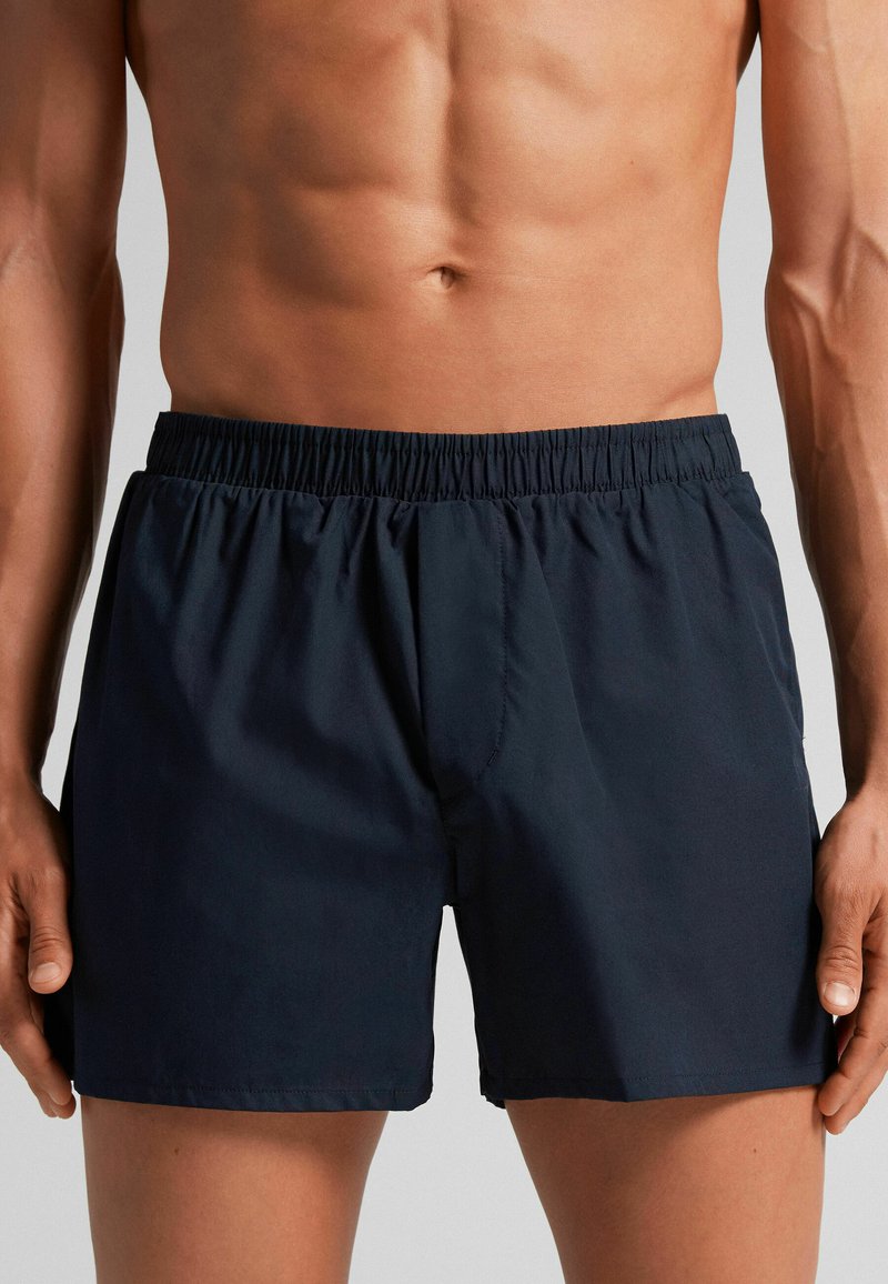 Námořnické modré plavky s elastickým pasem, hladkou texturou a mírně volným střihem. Žádné viditelné vzory ani detaily hardware.