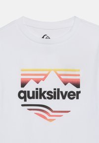 Λευκό μπλουζάκι με λογότυπο βουνού σε χρώματα ηλιοβασιλέματος πάνω από τη λέξη «quiksilver» με έντονα μαύρα γράμματα στο κέντρο του στήθους.