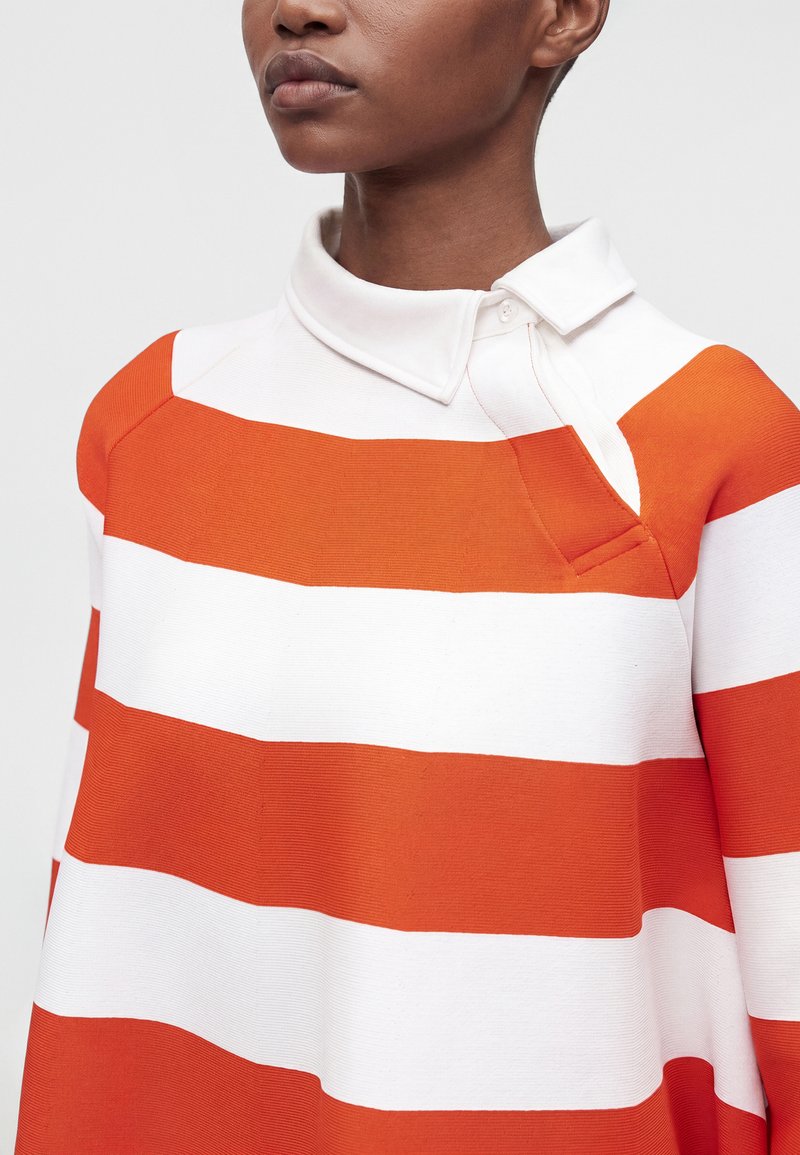 Personne portant une chemise à col avec de larges rayures horizontales orange et blanche, montrant le haut du torse et le cou sur un fond uni.