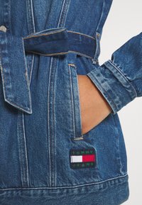 Chaqueta de mezclilla en azul oscuro con cintura ajustada, que presenta un parche etiquetado "TOMMY JEANS" en rojo, blanco y verde en el bolsillo.