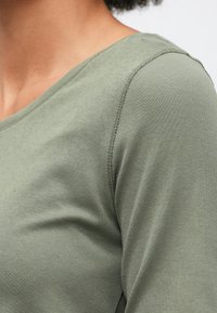 Κοντινό πλάνο ενός ατόμου που φοράει ένα ανοιχτό πράσινο, μακρύ μανίκι πουκάμισο με εμφανείς ραφές στους ώμους και στρογγυλή λαιμόκοψη.