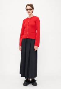 Cardigan en tricot rouge avec des boutons dorés, manches longues et une poche avant, associé à une jupe maxi noire fluide et des chaussures noires épaisses.