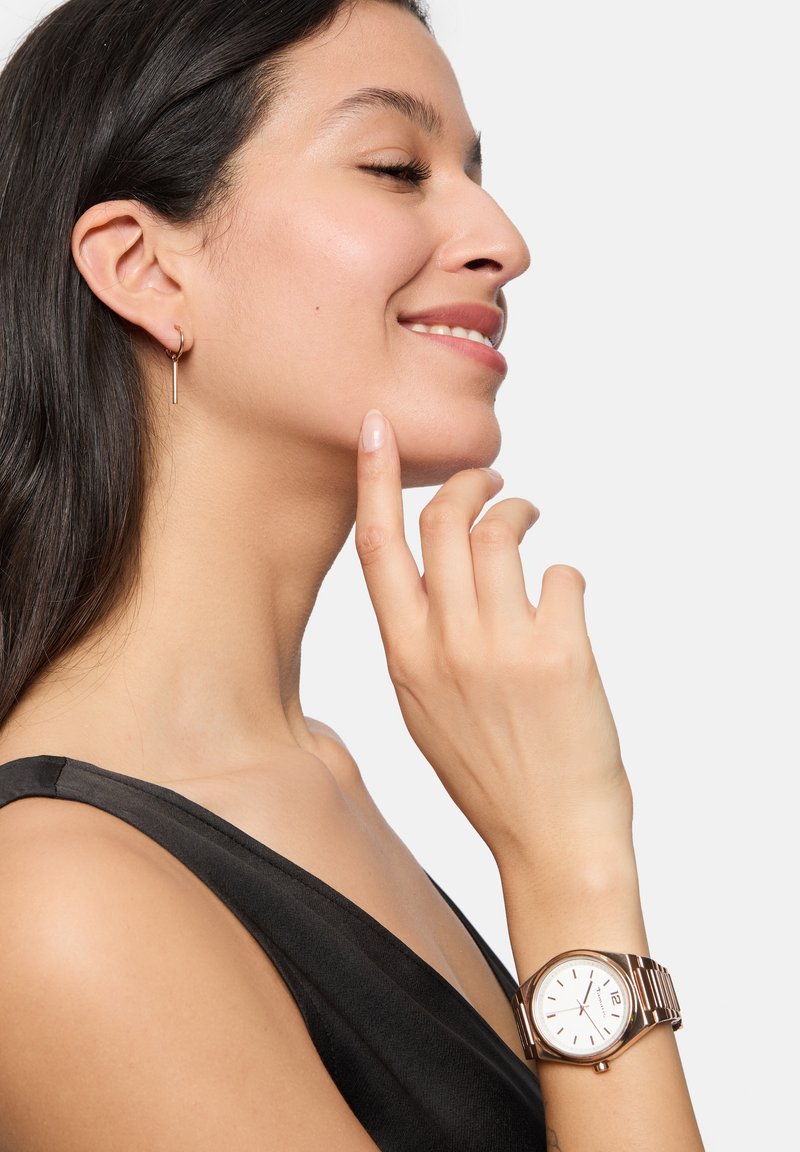 Rose-gouden polshorloge met een ronde witte wijzerplaat en zilveren uuraanduiders; model draagt een zwarte top, heeft lang donker haar en minimalistische oorbellen.