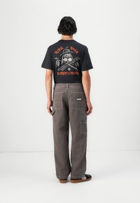 Schwarzes T-Shirt mit einem grafischen Design auf dem Rücken, kombiniert mit grauen, weit geschnittenen Cargo-Hosen, die mehrere Taschen und eine strukturierte Oberfläche aufweisen.