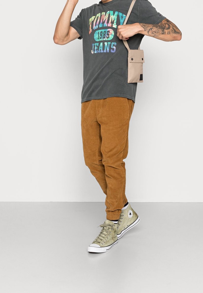 Bruna kordbyxor, grå grafisk t-shirt med "TOMMY JEANS 1985", beige axelremsväska och ljusgröna högskor.