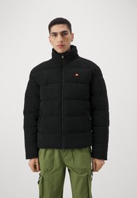 Schwarze Cord-Puffjacke mit hohem Kragen, Reißverschluss vorne und kleinem mehrfarbigen Logo auf der Brust. Strukturierter Stoff und gestepptes Design.