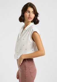 Camisa sin mangas blanca con cuello, adornada con pequeño bordado floral. Combinada con pantalones ajustados rosas, mostrando una vista lateral.