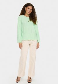 Zielona, plisowana bluzka z długim rękawem i okrągłym dekoltem, zestawiona z luźnymi, jasnobeżowymi spodniami i czarnymi sandałami na paski.