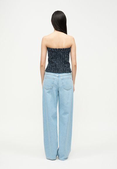 Femme debout, vue de dos, portant un haut sombre sans bretelles à motifs et un jean bleu clair à jambes larges, sur un fond blanc uni.