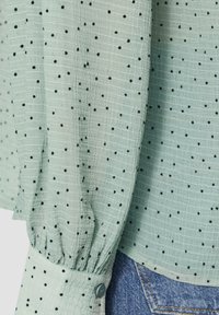 Jasnozielona bluzka w czarne kropki, z teksturowanej tkaniny i marszczonymi mankietami, z zapięciem na guziki przy nadgarstkach, w zestawie z dżinsami.