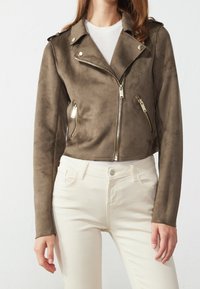 Donna che indossa una giacca da moto in suede marrone con cerniere argentate e pantaloni bianchi a vita alta, su uno sfondo bianco uniforme.
