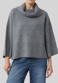 Femme portant un pull en tricot côtes gris ample avec des manches larges et un col châle large, associé à un jean en denim bleu.