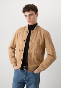 Tamnasti sudžučani bomber jakna s izrezanim ovratnikom, prednjim kopčanjem na gumbi i dva bočna džepa; nosi se preko crne rolke i traperica.