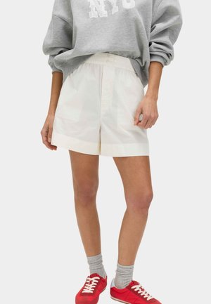 Person iført hvide højtaljede shorts, grå sweatshirt med "NYC"-tryk, grå sokker og røde sneakers, stående mod en ensfarvet baggrund.