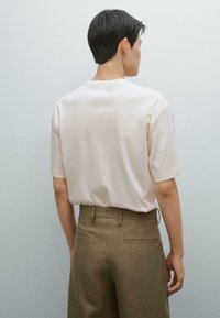 Camicia corta a maniche corte di colore beige chiaro, in cotone, con collo rotondo e vestibilità morbida, abbinata a pantaloni khaki con tasche posteriori.