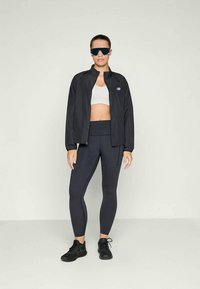 Ženska stoji oblečena v črno športno jakno, temne legice, belo športno modrček, črne športne copate in velike črne sončna očala pred enobarvno ozadjem.