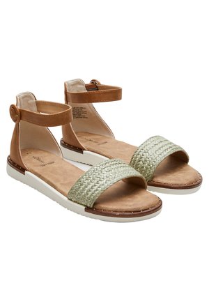 Par de sandalias planas para mujer con correas tobilleras de color caramelo, correas delanteras trenzadas beige, plantillas acolchadas y suelas de goma blancas.