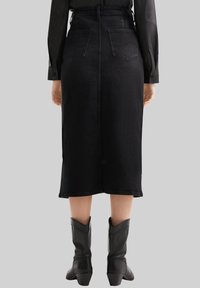 Vrouw in een zwarte midi rok met hoge taille en achterzakken, met zwarte laarzen tot halverwege de kuit, staand tegen een effen grijze achtergrond.